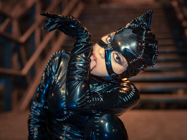 HannaSotoo in shiny black latex outfit, cat mask with ears, and gloves, kneeling, biting her finger, looking up with a seductive expression. Boobs, ass, and tits are covered.