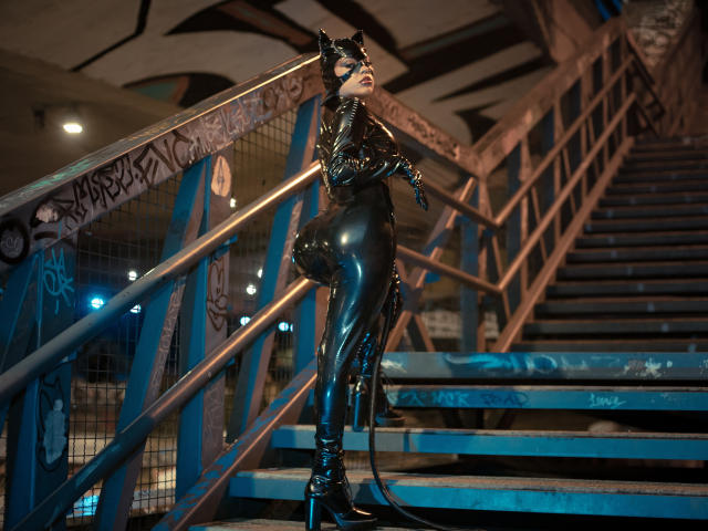 HannaSotoo in a shiny black latex cat suit with a mask, standing on blue stairs, looking back, with a long tail, wearing high heels, in a gritty, urban setting.