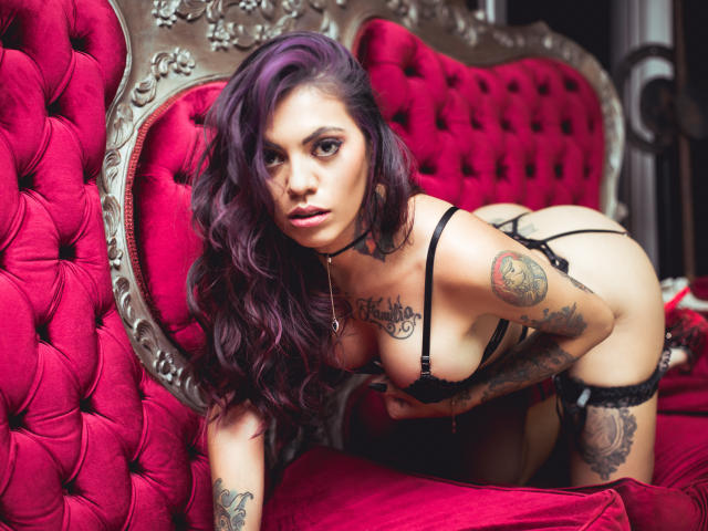 HannaSotoo with purple hair, tattoos, black lingerie, on red tufted sofa, on knees, looking at camera, showing tits, ass, and g-string. She's got a sexy, confident look.