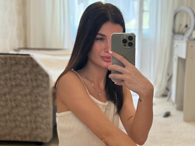 AmeliaSweetyX with long brown hair, wearing a white tank top, taking a mirror selfie with a gray iPhone, showing her tits and a cross necklace.