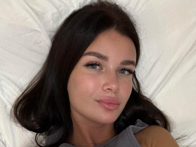 AmeliaSweetyX with dark brown hair, blue eyes, and pink lips, lying on a white pillow, wearing a gray top, showing a subtle smile.