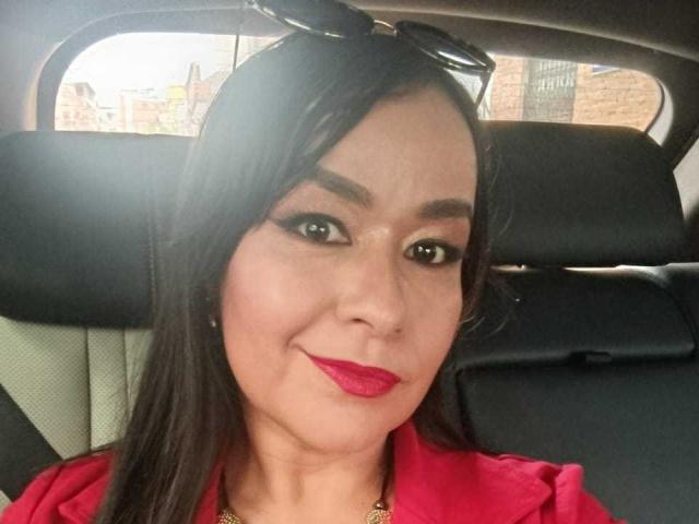 TamaraFoxy mit langen, schwarzen Haaren, rotem Lippenstift und dunklem Eyeliner, trägt ein rotes Hemd und sitzt in einem Auto. Sie hat eine Sonnenbrille auf dem Kopf und lächelt in die Kamera..