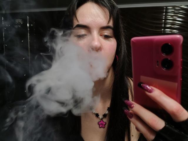 BerryDiva with long black hair, nose pierced, smoking, holding pink phone with glitter nails, wearing star necklace, taking selfie, white smoke in face, dark background.