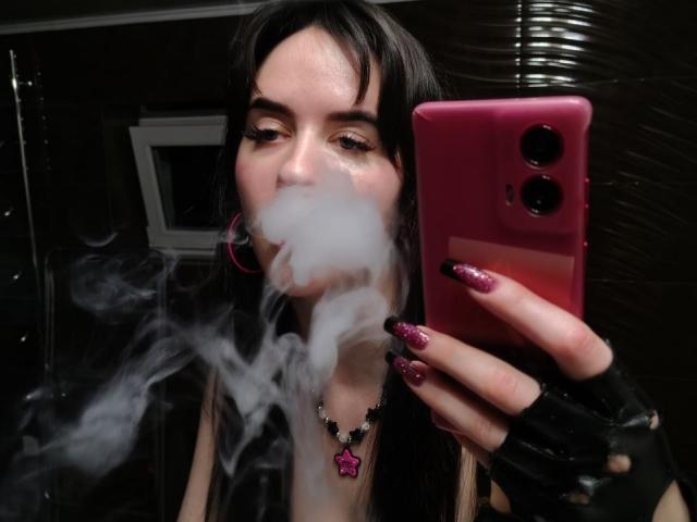 BerryDiva with black hair, holding pink phone, smoking white vapor, wearing pink hoop earrings, black fingerless gloves, and purple glitter nails, necklace with pink gem.