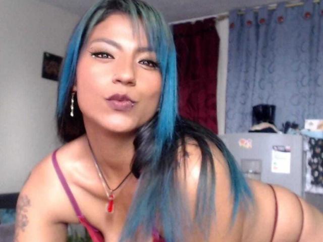 SamathaMay with blue and black hair, pouty lips, and a red necklace, wearing a pink bra, has a tattoo on her arm and is leaning forward, showing her ass.