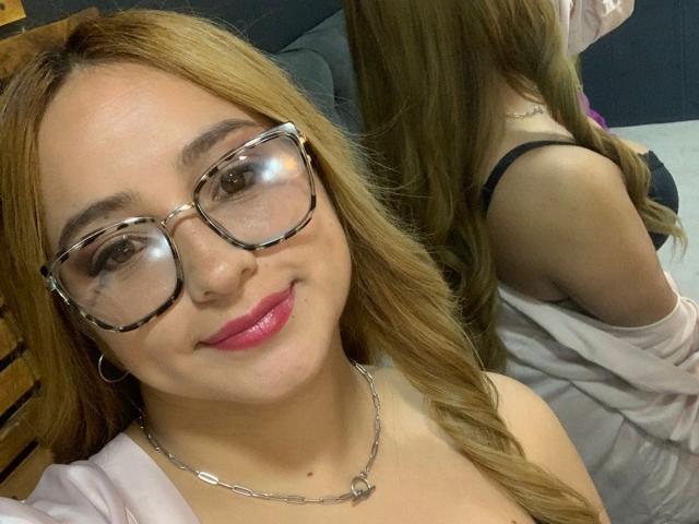 MaddyScarlett with long blonde hair, glasses, pink lipstick, and a necklace, smiling at the camera, showing her back with black bra straps and white shirt.
