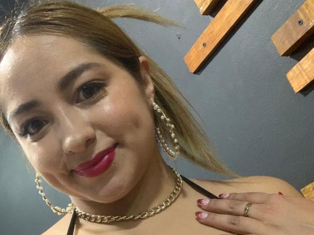 MaddyScarlett has light brown skin, red lipstick, and blonde hair in a ponytail. She's wearing gold chain earrings and a necklace, with her hand on her shoulder.