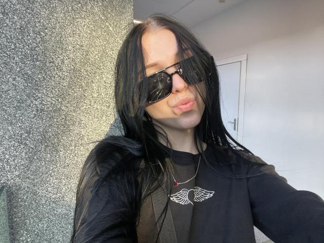 AmaiRin with long black hair, wearing black sunglasses and a black shirt with a wing design, pouting her lips, standing in front of a textured gray wall.