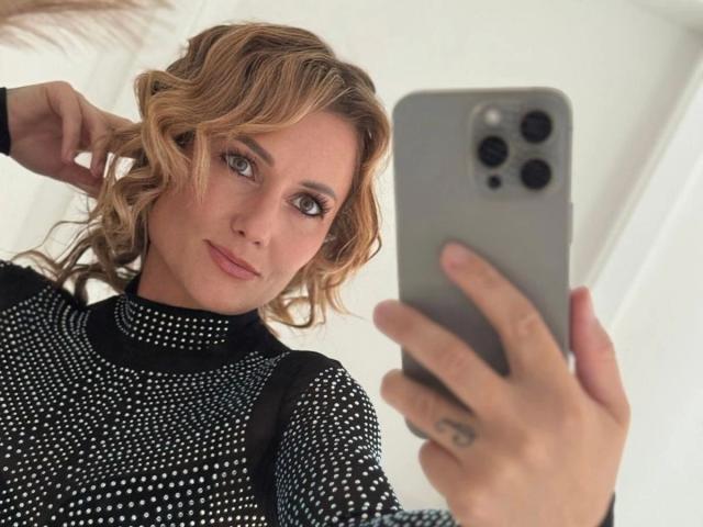 KylieFoxx-hot with curly blonde hair, wearing a black polka dot top, taking a selfie with a gray iPhone, holding it with tattooed fingers, smiling softly at the camera.