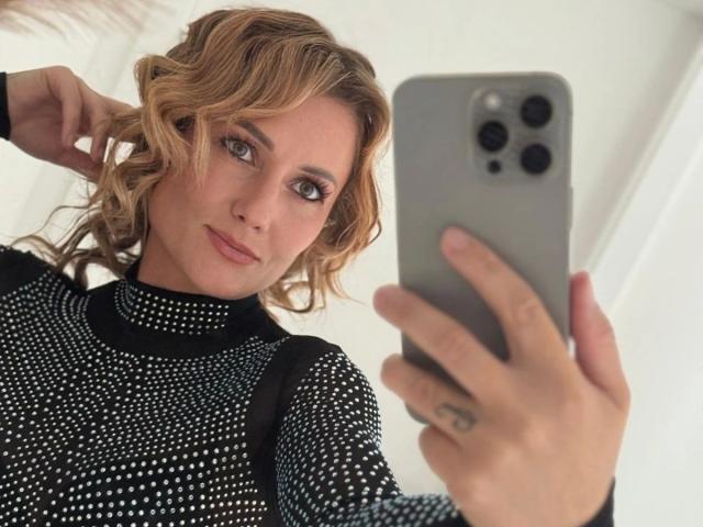KylieFoxx-hot with curly blonde hair, wearing a black polka-dot top, taking a selfie with a gray iPhone, looking at the camera with a slight smile.
