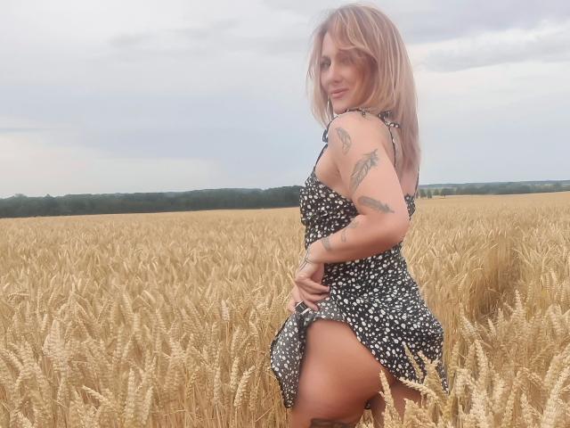 KylieFoxx-hot in a black, white-spotted dress, lifting it to show her bare ass, standing in a wheat field, with tattoos on her arms and a slight smile.