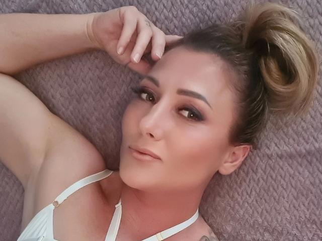 KylieFoxx-hot lying on a bed, looking at the camera with smoky eyes, wearing a white halter top, hand in hair, blonde bun, subtle makeup, pinkish skin, soft expression.