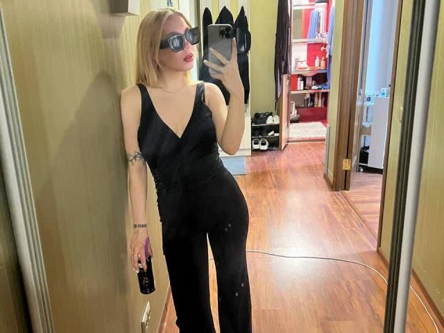 WildMaksi with blonde hair, glasses, black V-neck jumpsuit, holding phone and remote, tattoo on left arm, standing in front of mirror, wooden floor, mirror reflection.