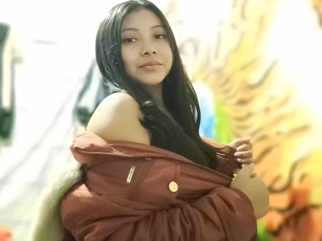 TammyGreyy with long black hair, wearing a red jacket off her shoulders, showing her black top, smiling subtly, holding jacket buttons, in a blurred indoor setting.