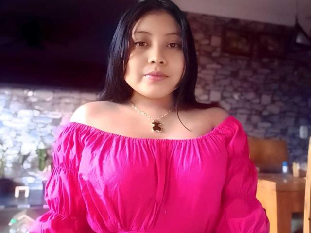 TammyGreyy with long black hair, pink off-shoulder top, small tits, and a cute necklace with a butterfly charm, looking slightly to the side, smiling softly.