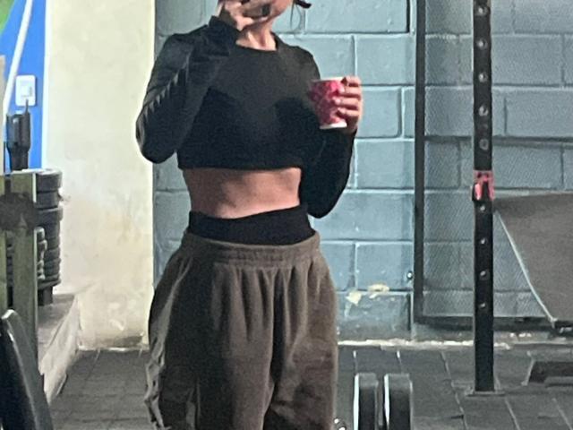 SteffanyTaylor in a gym, wearing a black long-sleeve crop top and loose brown pants. She holds a red cup in her right hand, with her left hand near her mouth. Her midsection is exposed. The background includes a blue wall, a weight rack, and a metal pole. The image has a casual, realistic style. The lighting is natural.