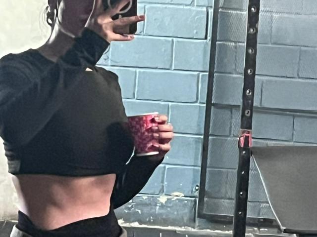 SteffanyTaylor with her tits showing in a black sports bra, holding a red fruit, mirror selfie in a gym, right arm raised, black hair, tight black pants.