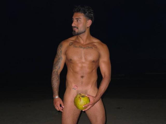 Naked IvannaAndFriend with a muscular build stands against a dark background. He holds a green coconut with his right hand, covering his genitals. He has tattoos on his left arm and chest, a trimmed beard, and short, dark hair. His left wrist has a silver bracelet. He looks to the left, showing a confident expression. His skin tone is tan. The image focuses on his nudity and the coconut. The composition emphasizes his physical fitness and tattoos.