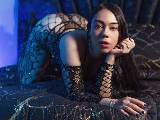 Tahira Thompson, con lunghi capelli neri, indossa un abito di pizzo nero, è a quattro zampe, mostra il sedere, su un letto con luci blu e dorate, e guarda la fotocamera..