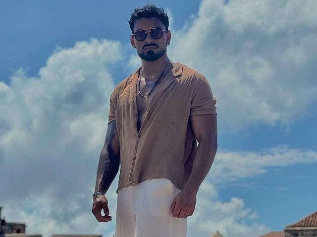 IvannaAndFriend is a muscular dude with dark hair and beard, wearing a tan shirt and white pants, sporting sunglasses and tattoos, standing against a blue sky with clouds.