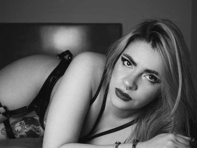 Fotografia in bianco e nero di Alissa Spellman con lunghi capelli biondi, rossetto scuro, piercing al naso e lingerie nera, sdraiata sul letto, che mostra un tatuaggio sulla coscia destra, mentre guarda verso la fotocamera..