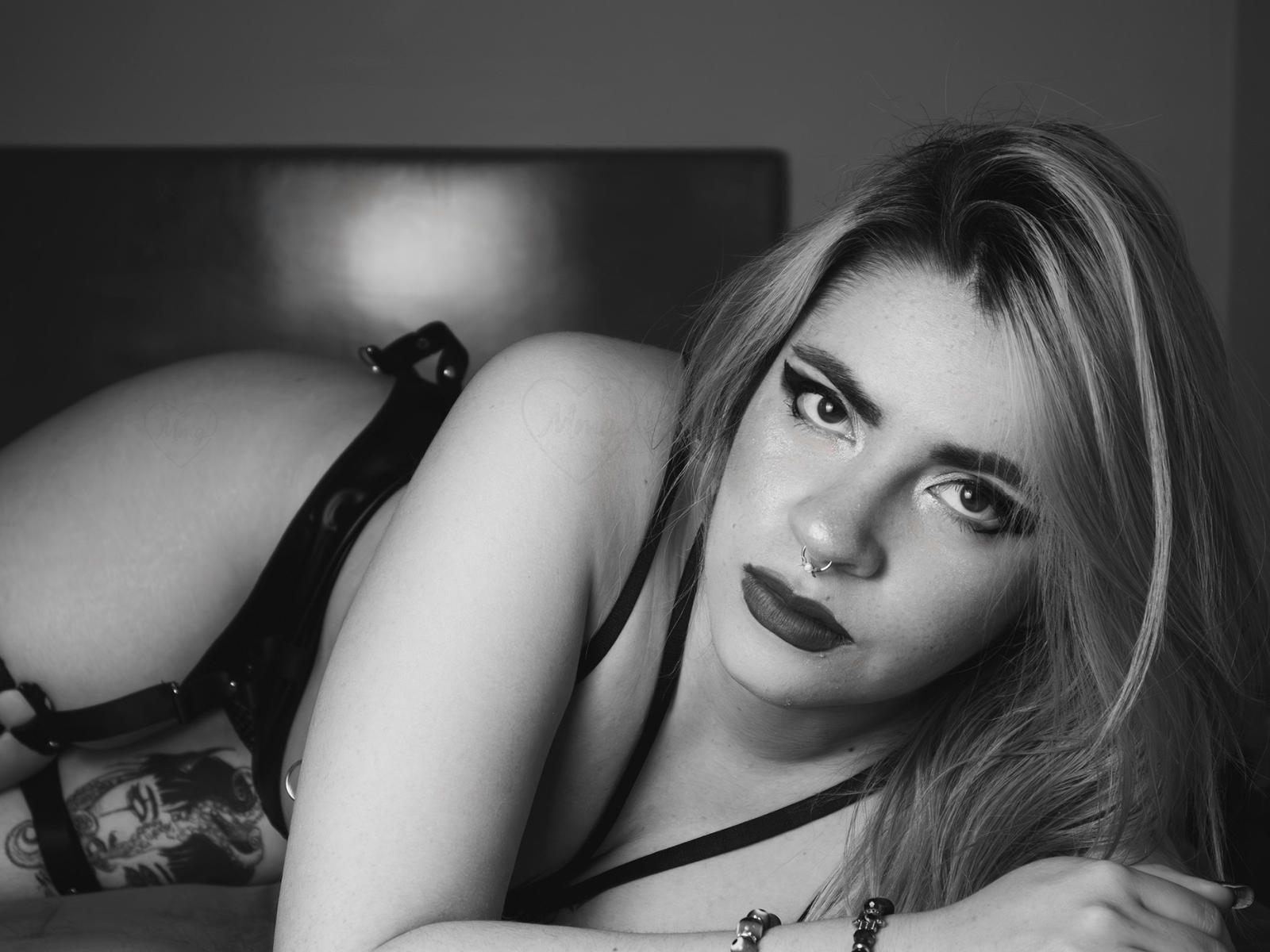 Black-and-white photo of AlissaSpellman with long blonde hair, dark lipstick, nose ring, and black lingerie, lying on bed, showing tattoo on right thigh, looking at camera.