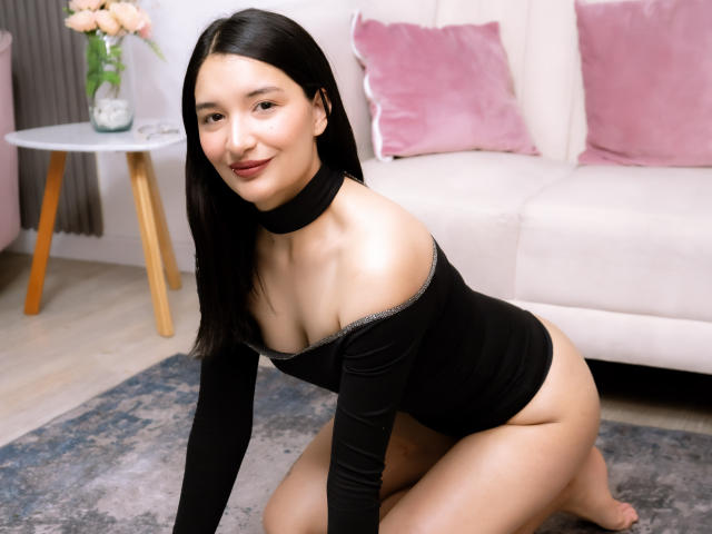 HaileyMonsalve with black choker, black off-shoulder top, and black bottom, kneeling, smiling, showing her ass and small tits, with straight black hair, in a living room.