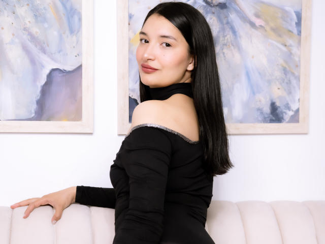 HaileyMonsalve with black hair, off-shoulder black top, sitting on white couch, looking over shoulder, smiling slightly, hand on couch, abstract paintings in background, soft lighting, casual.