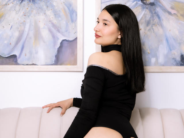 HaileyMonsalve with black hair, black off-shoulder top, and bare ass, sitting on white couch, looking back, smiling, white wall with blue abstract paintings in background.