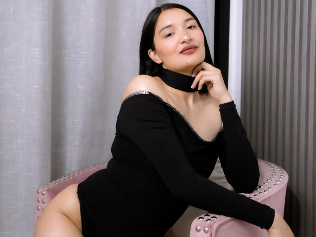 HaileyMonsalve with black dress, bare shoulders, choker, sitting on pink chair, hand near face, smiling, dark straight hair, light skin, small tits, cute, sexy, confident.