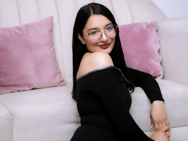 HaileyMonsalve with long black hair, glasses, and a black off-shoulder top, smiling at the camera, sitting on a white couch with pink pillows, showing her bare shoulder.