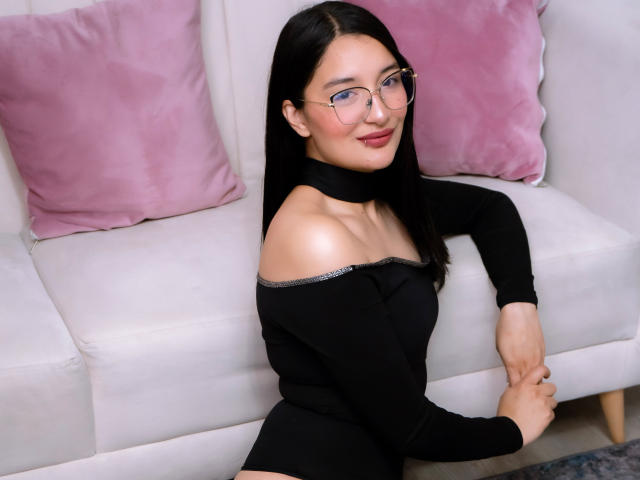 HaileyMonsalve with long black hair, glasses, and a black off-shoulder top, sitting on a white sofa with pink pillows, smiling, showing her bare shoulders and hands.