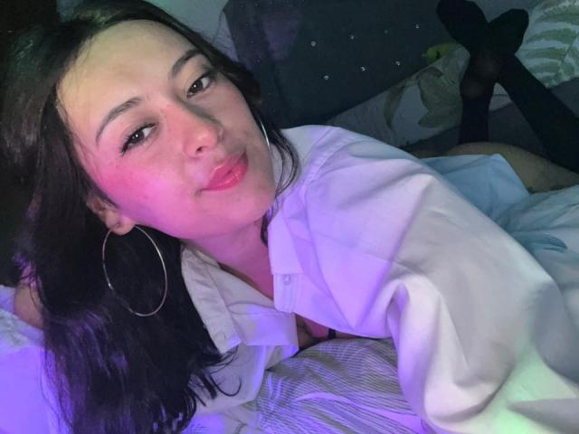 DamiAmbani with long black hair, pinkish glow on her face, wearing a white shirt, hoop earrings, and a slight smile, lying on a bed with her legs up.
