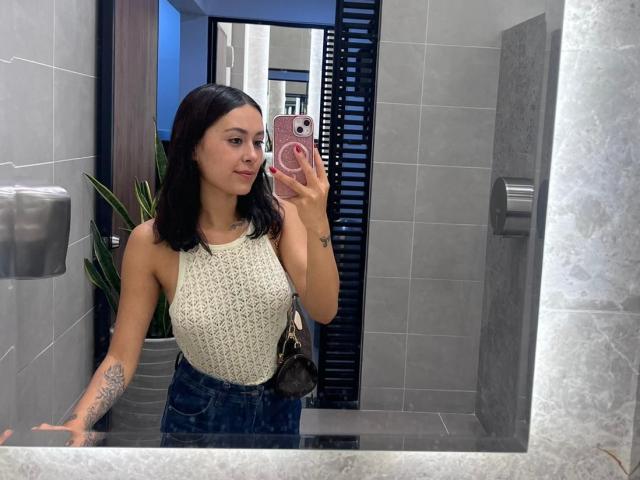 DamiAmbani with dark hair, white lace sleeveless top, blue jeans, small tattoo on right arm, holding pink phone, mirror selfie in gray-tiled bathroom, small black purse.