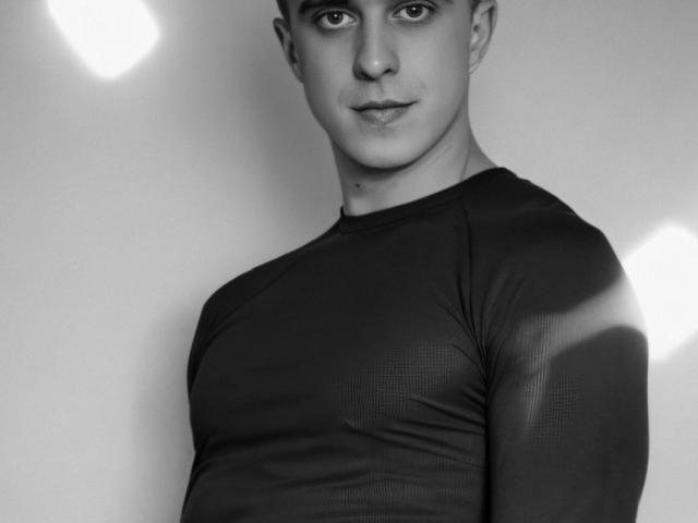 JhonShelby with a strong, handsome face, wearing a tight, black, long-sleeve shirt, showing a muscular chest, and a slight, confident smile. Black and white, shadowed.