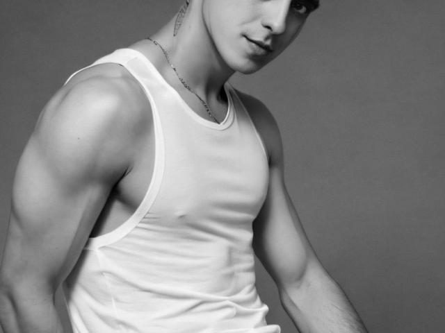 JhonShelby's muscular upper body in a white tank top, showing defined arms and chest, with a slight smile and short hair, wearing a thin necklace. Black and white.
