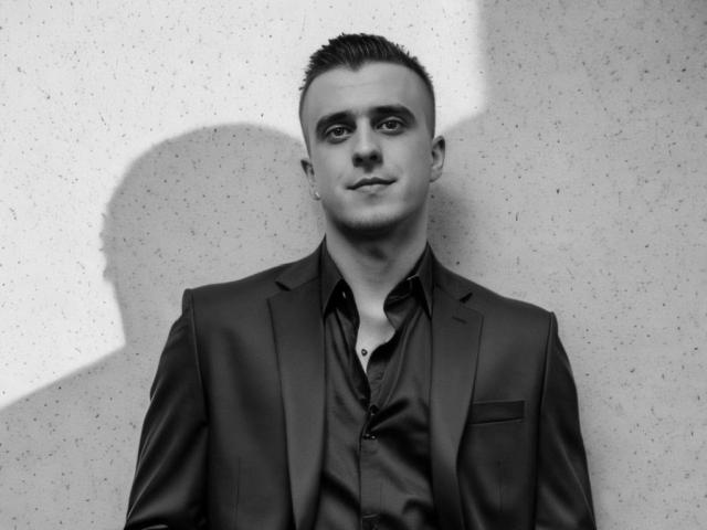 Black and white photo of JhonShelby with short, slicked-back hair, wearing a dark suit and shiny shirt, shadow on the wall, looking slightly to the left.