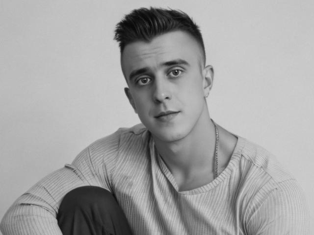 JhonShelby with short, spiked hair, wearing a striped shirt, sitting with a slight smile, looking directly at the camera, and a thin chain necklace. Black and white photo.
