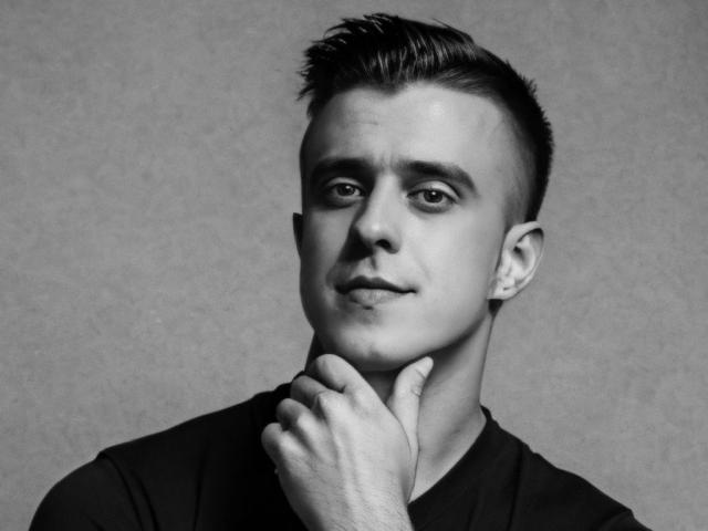 JhonShelby has short, dark, spiked hair, a clean-shaven face, and is touching his chin with a slight smirk, wearing a black shirt in a grayscale photo.
