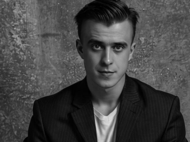 JhonShelby, a young dude with a slicked-back haircut, wearing a pinstripe suit and white tee, has a serious face and a clean-shaven look. Black and white photo.