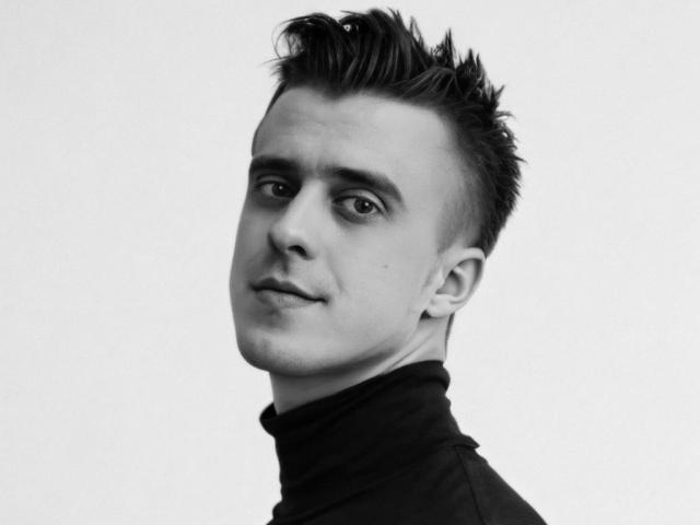 Black and white photo of JhonShelby with short, spiked dark hair, a turtleneck, and a slight, confident smile. His eyes are focused and his expression is calm.