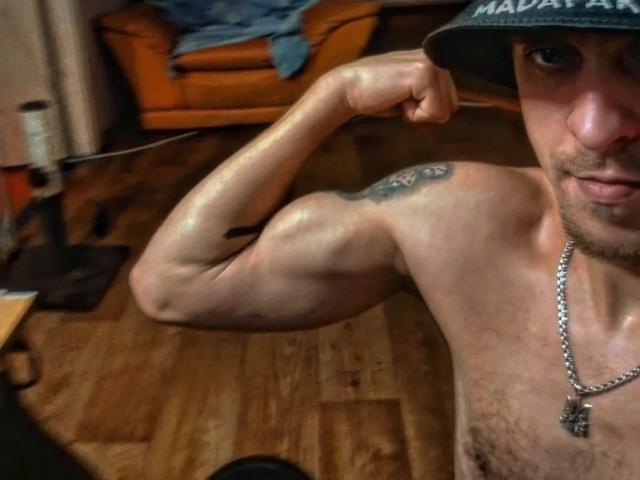 JhonShelby, shirtless, flexing bicep, visible tattoo on shoulder, wearing baseball cap, necklace with cross, wooden floor, slight chest hair, looking at camera, in dimly lit room.