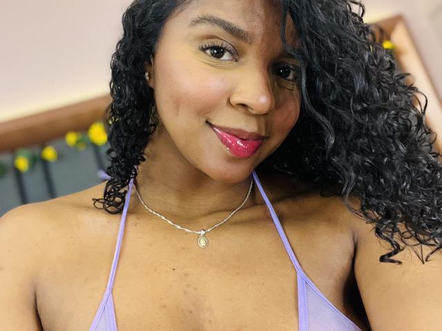 LisaMendez, met krullend zwart haar, bruine huid, roze lippenstift en een zilveren ketting, draagt een paars topje met halter en glimlacht zachtjes. Haar borsten zijn zichtbaar, waardoor een lichte decolleté te zien is..