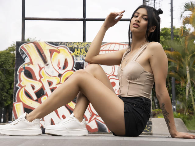 AlisKooper with tan bralette, black shorts, white sneakers, sitting on concrete, legs crossed, right arm raised, tattoo on left arm, black hair in bun, green eyes, medium tits.