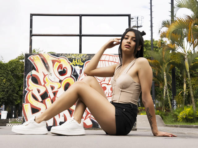 AlisKooper with tan bralette, black shorts, white sneakers, sitting on pavement, legs crossed, hand on head, palm tree, graffiti, tattoo on arm, sexy, tan skin, dark hair, confident.