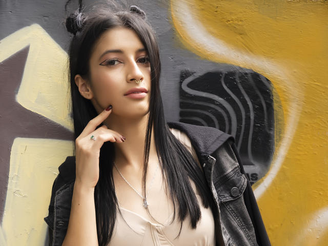 AlisKooper with long black hair, nose ring, and eyeliner, wearing a black jacket over a beige top, touching her chin, looking to the side, with a ring on her finger.