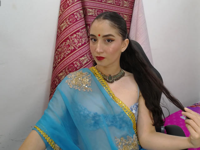 JalimaMona with dark hair in a ponytail, wearing a blue and gold sheer sari, red lipstick, and a red bindi, holding her hair, with a black necklace.