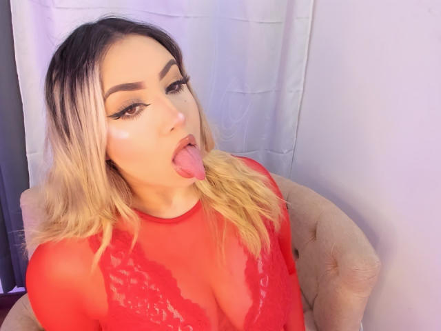 MelissaLust with blonde hair, red lace top, and thick eyeliner, sticking out her tongue, showing off her shiny, painted-up face and red sheer top that covers her tits.
