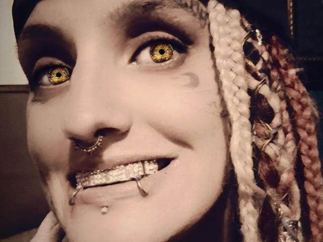 SexySavageNova with yellow, spiral-patterned eyes, nose ring, and metal-studded grin showing multiple teeth and a bottom lip piercing. Pink and white braided hair on the right.