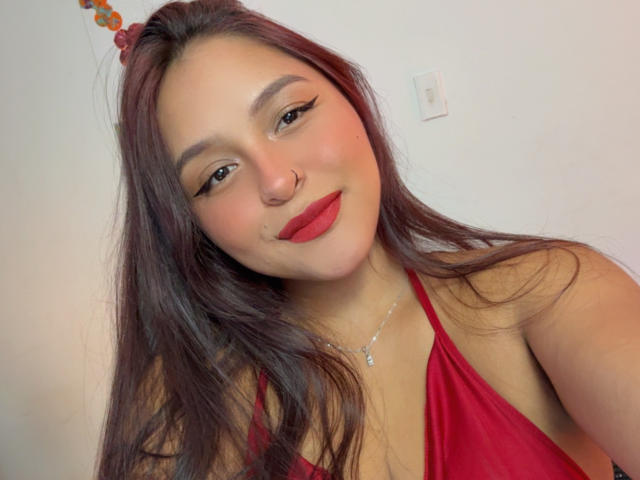 MarianaVargasX has long brown hair, red lipstick, winged eyeliner, a nose ring, and is wearing a red halter top. Her ass is not visible.